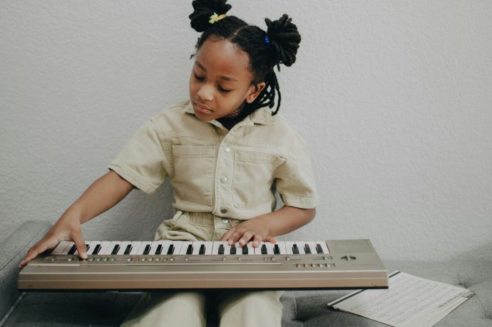 A young girl with dreadlocks playing an electronic keyboard on a gray sofa, creating a serene and minimalistic scene.